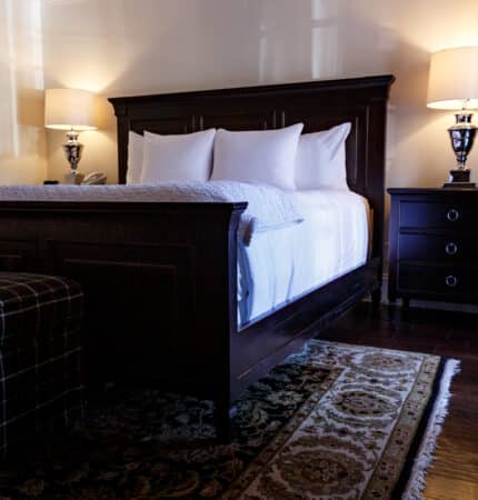 A neatly arranged bedroom with a dark wooden bed frame, white bedding, a patterned rug, two bedside tables, and two lit lamps. A checkered bench is placed at the foot of the bed.