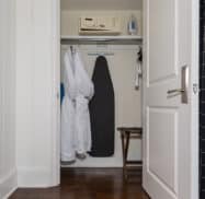 An open closet with two white robes, an ironing board, an iron, a shelf, and a small table. A thermostat is mounted on the wall outside the closet. The closet door is partially open.