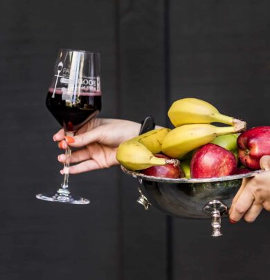 A person holds a glass of red wine and a silver bowl containing bananas and apples.