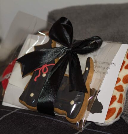 Gift with a black bow containing a cookie shaped like a dress, a card, and an item with orange spots, placed on a gray surface.