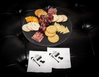A platter with assorted crackers, cheese slices, salami, and grapes on a black surface. Two branded napkins are placed in front.