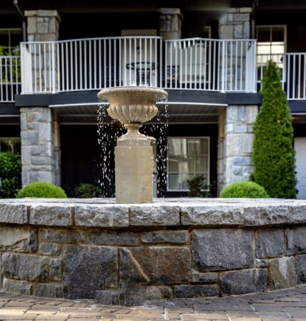 Stone fountain with cascading water in front of a building with balconies and two lampposts. Surrounded by greenery.