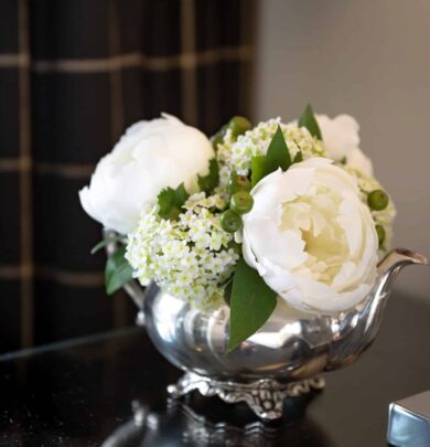 A silver vase holds white peonies and small white flowers with green leaves, placed on a reflective surface. A dark plaid curtain is partially visible in the background.