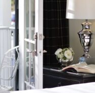 A bedside table with a lamp, an open book, and a bowl of white flowers next to an open glass door leading to a white chair outside.