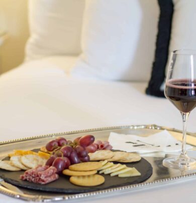 A silver tray with crackers, cheese, grapes, and salami next to a glass of red wine on a bed.