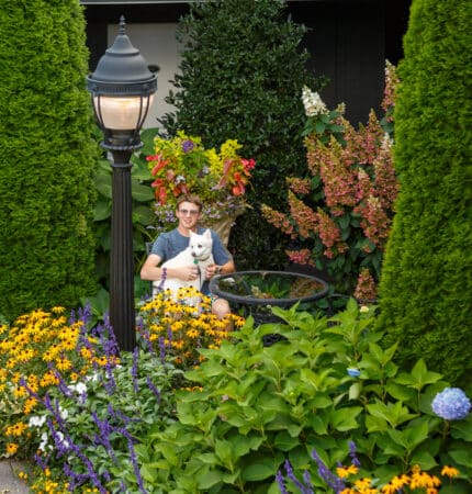 A guest at Highlands Suites Hotel relaxes on a patio chair, cradling a dog amidst the garden's lush greenery, vibrant flowers, and an elegant lamppost.