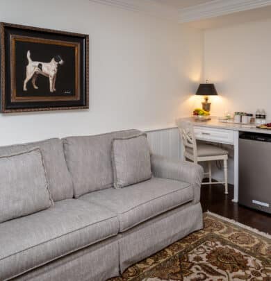 Cozy living area with a gray sofa, framed dog painting, small desk with a lamp, mini fridge, and an area rug.