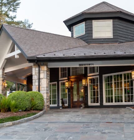 A modern building with large windows, stone accents, and a sloped roof. Surrounded by shrubs, the entrance features a paved walkway and overhead lighting.