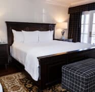 A neatly made bed with a white quilt and pillows is flanked by two lamps on dark wooden nightstands in a bedroom with hardwood floors and a checkered ottoman.