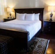 A neatly made bed with white linens and two bedside tables, each holding a lamp, in a room with dark wood furniture and a patterned rug.