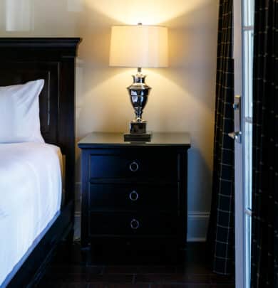 Bedroom interior with a dark wooden nightstand, lit lamp, and adjacent bed with white bedding. Dark curtains are partially drawn near a doorway.
