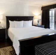 A bedroom with a large dark wood bed, white bedding, two bedside tables with lamps, a window with dark curtains, and a checkered ottoman.