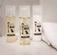 Three small hotel toiletries include body wash, shampoo, and conditioner on a white surface next to folded towels.