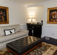 Living room with a beige sofa, a large ottoman, and two dog portraits on the wall. A small desk with a lamp and chair is in the corner. A pet bed is on the floor.