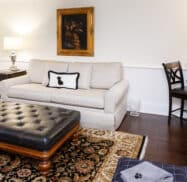 A cozy living room with a beige sofa, black leather ottoman, table lamp, and a black chair at a desk. A framed dog portrait hangs above the sofa. A patterned rug covers the wooden floor.