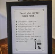 Framed sign on a wooden table listing various items for sale, including wine glasses, dog treats, lotions, towels, and an umbrella, with prices.