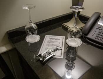 A hotel room desk with two upside-down wine glasses, a corkscrew, a telephone, and a menu labeled "libations.