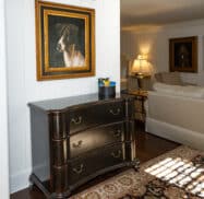 Room with a wooden chest, dog portraits on walls, beige sofa, and a table lamp casting light.