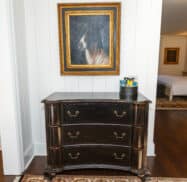 A dark wooden dresser with three drawers stands beneath a framed portrait of a horse. To the right, a small round container holds assorted snacks. Doorways are visible on either side of the dresser.