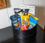 A black container holds Planters cashews, M&M's, Snickers, Pringles, Oreos, and Twizzlers. A sign behind reads "Enjoy Now...Pay Later.