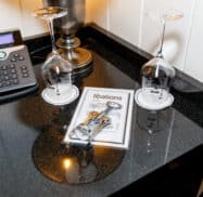 Two upside-down wine glasses, a corkscrew, and a "libations" menu on a black countertop beside a phone and a lamp.