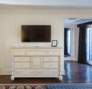 Room interior showing a white dresser with a TV mounted above, flanked by French doors with dark curtains and wooden floors.