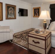 Living room with a beige sofa, plaid armchair, and trunk-style side table. Three dog portraits hang on the walls. A TV is mounted opposite the window with plaid curtains.