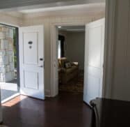 A hallway with open doors leading to a sunlit exterior and a room with a beige couch. Wood flooring and white walls are visible.
