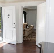 Open doors reveal a bright living room with a beige sofa and patterned rug, visible from a hallway with white walls and wooden flooring.