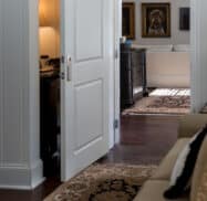 A cozy living room with a partially open door, revealing a lamp-lit room. The area features patterned rugs, paintings on the wall, and a beige sofa.