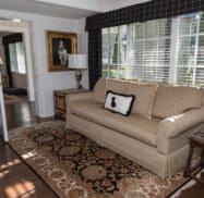A living room features a beige sofa with a cushion and framed dog portrait, a patterned rug, wooden floors, and windows with blinds. An open door leads to another room.