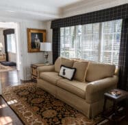 A cozy living room with a beige sofa, a patterned rug, and a side table. Large windows with dark curtains let in light. A framed painting and a lamp are in the background.