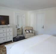 A cozy bedroom reminiscent of the best hotels in Highlands, NC, featuring a large bed, white dresser, wall-mounted TV, armchair, and an intricately detailed carpet.