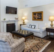 Living room with a white sofa, plaid chairs, a black ottoman, two lamps, a fireplace, and a wall-mounted TV. A framed painting hangs above the sofa.