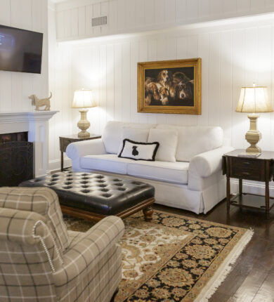 Cozy living room with a white sofa, plaid armchair, and dark leather ottoman. A painting and a wall-mounted TV are above the sofa. Two lamps and a fireplace add warmth.