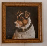 A framed painting of a brown and white dog wearing a collar is hung on a white wall.