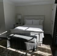 Bright bedroom with a neatly made bed, two side tables, a lamp, and a bench at the foot. Sunlight filters through the window, casting shadows on the walls and floor. Experience this serene retreat in one of the best hotels in Highlands NC.