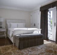 A neatly arranged bedroom with a white bed, bedside table, plaid bench, and patterned rug. Sunlight filters through a window with plaid curtains, offering the perfect retreat for those seeking lodging in Highlands NC.