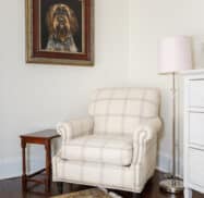 A white armchair with plaid design next to a wooden side table and lamp. A framed dog portrait hangs on the wall above the chair. A patterned rug covers the dark hardwood floor.