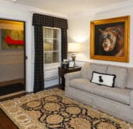 A cozy living room with a couch, plaid armchair, and a decorative rug. A portrait of a bear hangs above the couch, and a lamp sits on a side table. A door is partially open.