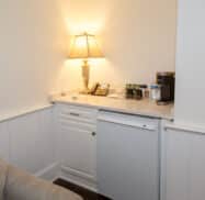 A small cozy corner with a lamp, telephone, coffee maker, and bottled water on a countertop above a white mini fridge next to a beige sofa.