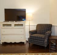 A room with a black plaid armchair, a small wooden side table, a cream dresser with a flat-screen TV, and a tall floor lamp. A pet bowl on a mat is near the dresser.