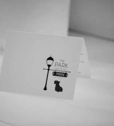 A black and white card artfully captures a streetlamp with a "The Park On Main" sign, featuring the silhouette of a small dog nearby—a charming scene reminiscent of downtown near the Highlands NC hotel.
