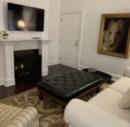 Living room with a wall-mounted TV, a fireplace, a leather ottoman, a white couch, and a large horse painting on a white paneled wall.