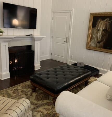 Living room with a wall-mounted TV, a fireplace, a leather ottoman, a white couch, and a large horse painting on a white paneled wall.