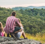 A man and two children sit on a rocky hill, overlooking a scenic view of green forests and distant mountains.