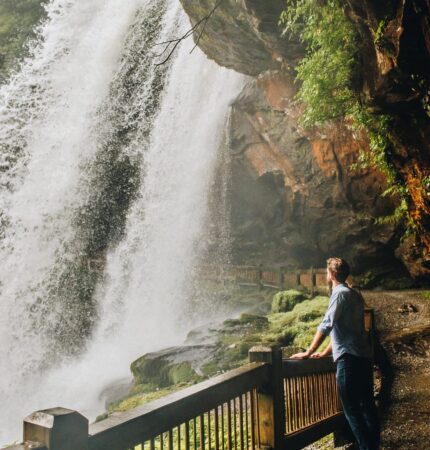 A person stands on a path beside a large, flowing waterfall surrounded by greenery and rocky cliffs.