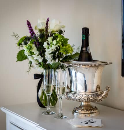 Silver ice bucket with champagne, two glasses, and a bouquet of white and purple flowers on a white dresser.