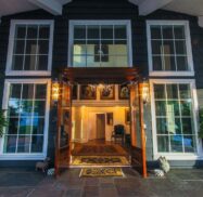 Entrance of a building with large glass doors, flanked by tall windows and decorative plants in urns. Inside, a chandelier and framed artwork are visible. A chair is positioned near the doorway.