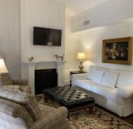 Living room with white couch, plaid chairs, patterned rug, dark ottoman, and wall-mounted TV above a fireplace. Paintings of dogs are on both walls. Classic decor with table lamps.